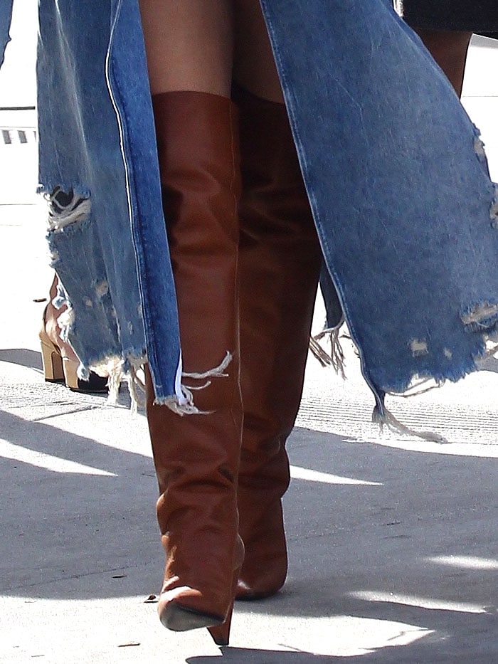 Close-up of Chrissy Teigen’s luxe Saint Laurent “Niki” thigh-high boots in rich cognac leather, styled with a distressed denim trench for an effortlessly chic street-style moment