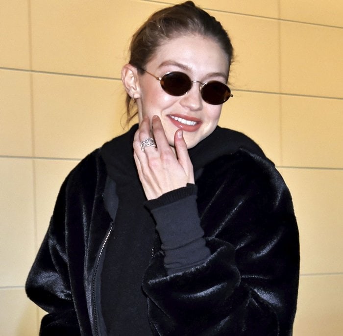 Gigi Hadid smiles at the camera, wearing sleek round sunglasses, a diamond ring, and a black velvet bomber jacket
