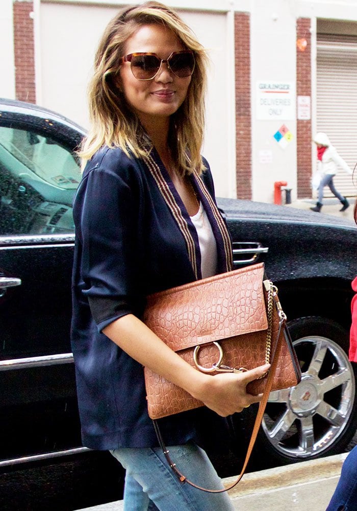 Chrissy Teigen elevates her casual jeans-and-blazer look with a blush-pink crocodile-embossed Coach clutch, blending effortless daywear with polished sophistication