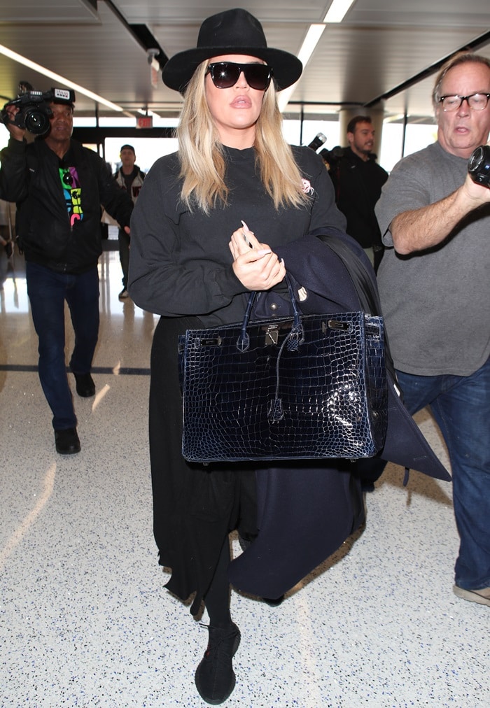 Khloé Kardashian sported a black pull-over sweatshirt, a wide-brim hat, black workout leggings, one more sweatshirt tied around her waist, designer sunglasses, and a long black trench coat