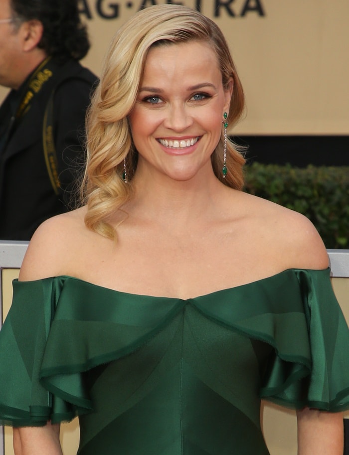Reese Witherspoon radiates elegance with soft waves, glowing skin, and dazzling emerald drop earrings that perfectly complement her off-the-shoulder gown