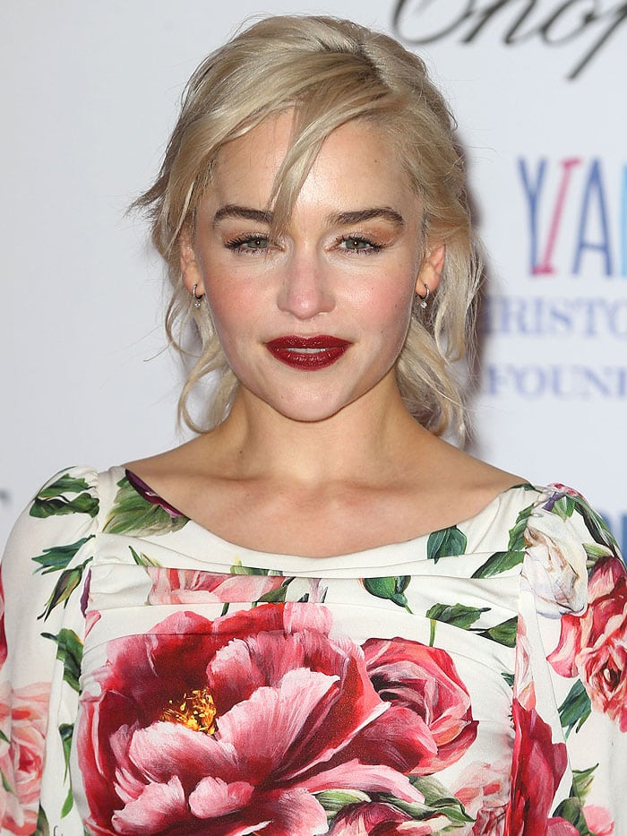 Emilia Clarke complements her Daenerys-inspired blonde locks with bold red lipstick and a soft, romantic updo, adding elegance to her floral Dolce Gabbana dress