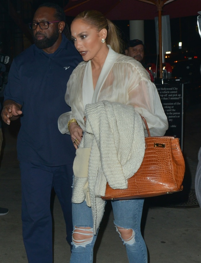 Balancing off-duty denim with luxury polish, Lopez layers a gauzy white blouse over ripped skinny jeans while carrying a Hermès shiny crocodile Birkin in the warm “Miel” shade