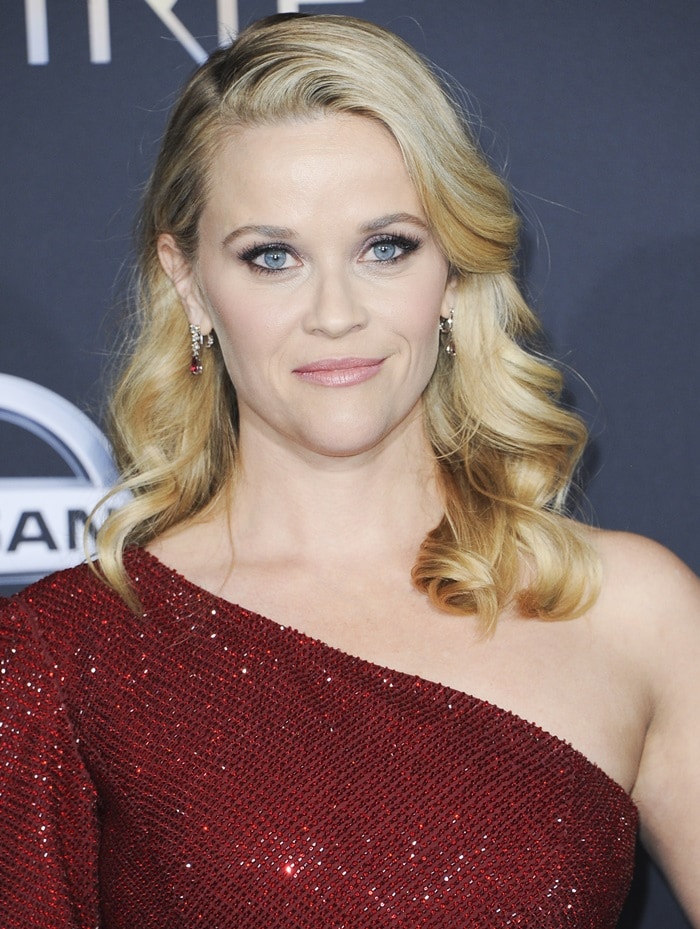 Reese Witherspoon’s softly waved blonde hair, smoky eye makeup, and ruby drop earrings complement the rich claret tone of her one-shoulder gown