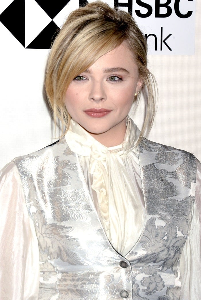 Chloë Grace Moretz paired a romantic white tie-neck blouse with dramatic bishop sleeves and a shimmering silver floral jacquard vest, blending Victorian-inspired elegance with the fashion-forward styling of Louis Vuitton's Spring 2018 collection