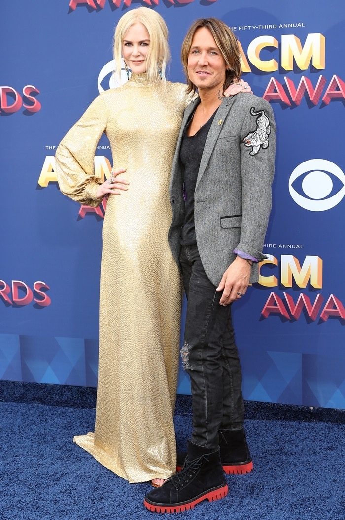 Nicole Kidman shimmers in a gold Michael Kors Collection sequin gown paired with matching metallic sandals, while Keith Urban complements her polished glamour in a grey embroidered blazer, black tee, distressed jeans, and rugged black boots with bold red soles