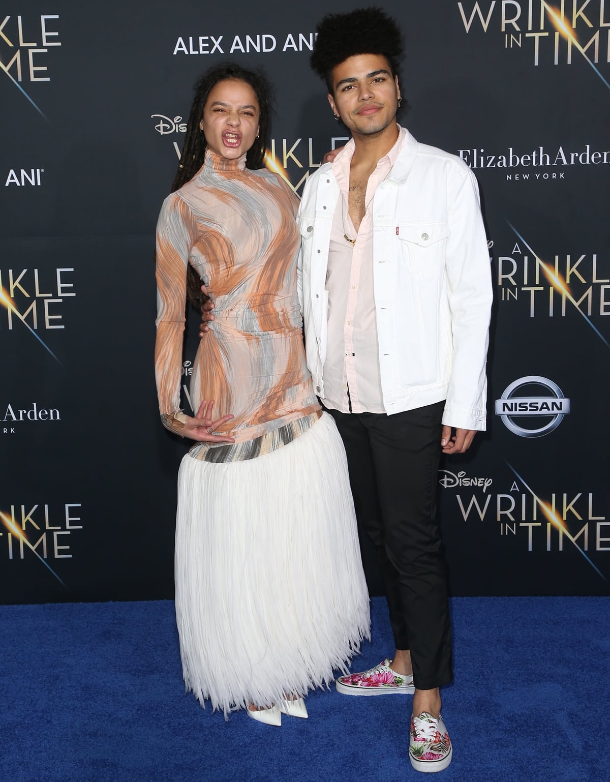A playful Sasha Lane posed with her brother, Sergio Darcy Lane, at the A Wrinkle in Time premiere, showcasing a Celine Spring 2018 gown with a marbled high-neck bodice, a flowing white fringed skirt, and sharp white Christian Louboutin pumps peeking out beneath the hem