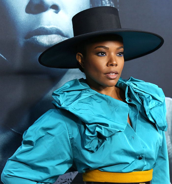 Gabrielle Union stuns in a bold turquoise ruffled blouse, oversized wide-brim hat, and striking electric blue eyeliner for a dramatic red carpet look