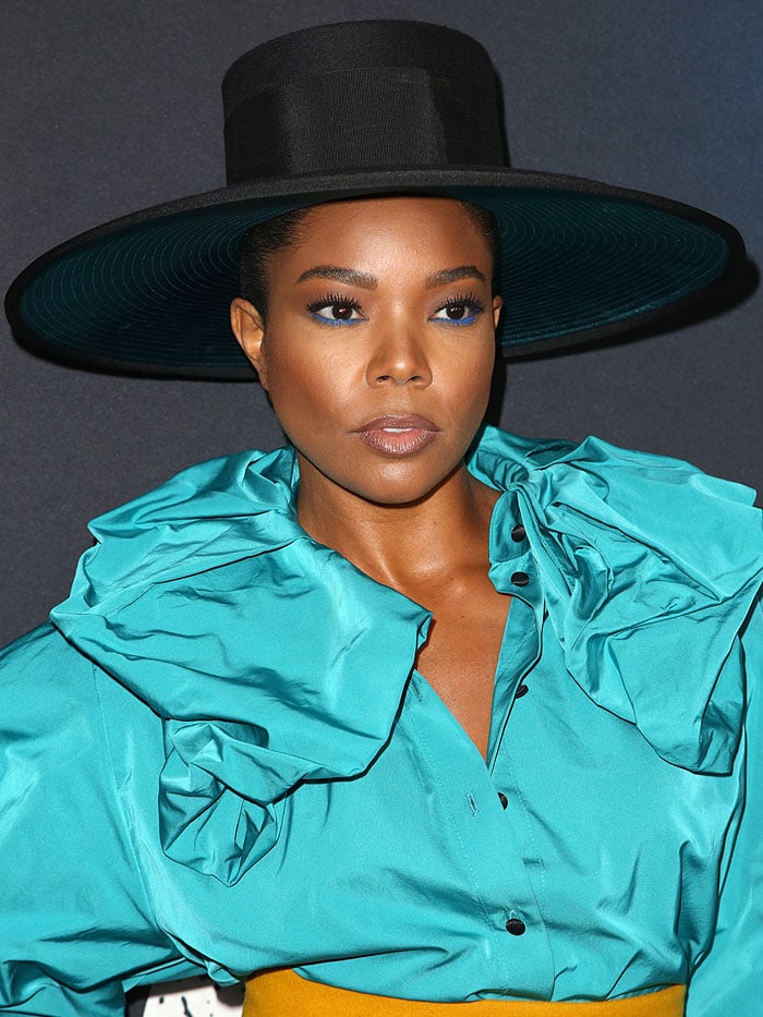 The blouse features voluminous ruffled detailing cascading down the shoulders and neckline, while Gabrielle’s glossy nude lips add a subtle contrast to her bold turquoise look