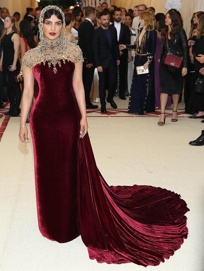 Priyanka Chopra rocked a Ralph Lauren Collection burgundy velvet column gown with a long train and a jeweled hood headpiece at the 2018 Met Gala