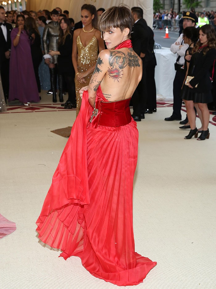 Ruby Rose wearing a Tommy Hilfiger red pleated dress with a velvet sash, gold Jimmy Choo Kaylee sandals, and dangling gold crucifix eyebrow jewelry at the 2018 Met Gala
