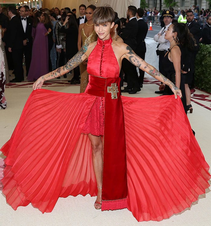 Ruby Rose shows off her tattooed legs in a pleated red Tommy Hilfiger gown with a high slit and priestly sash, finishing the look with gold Jimmy Choo "Kaylee" sandals and signature ink on full display