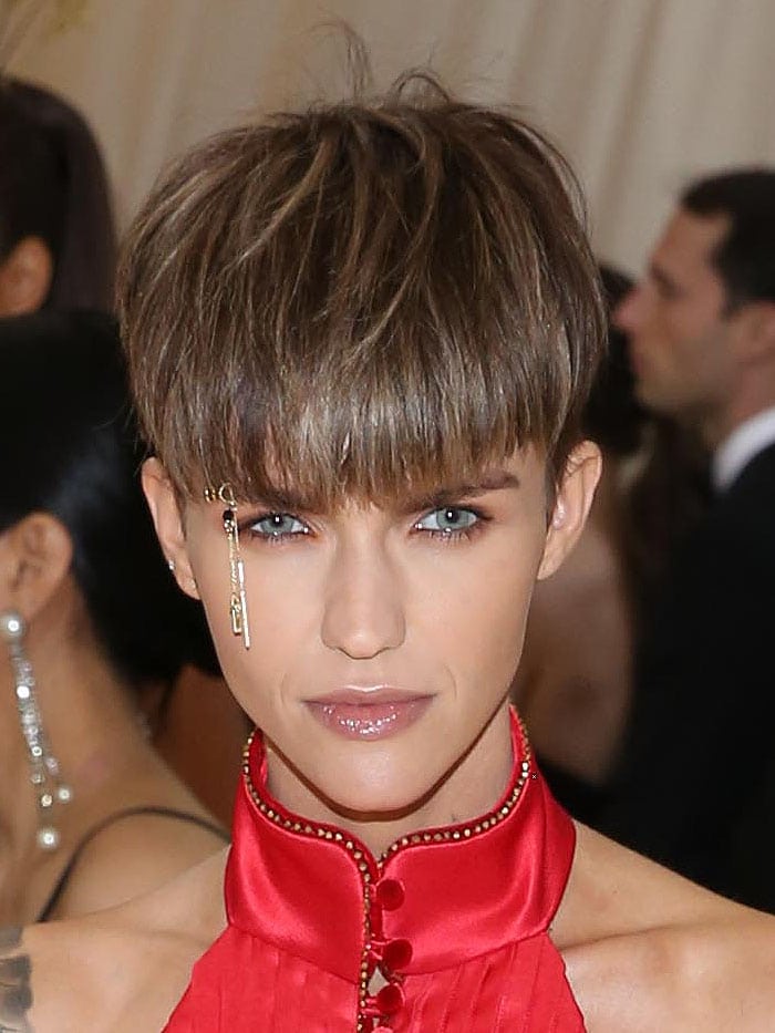 Ruby Rose puts an edgy twist on Met Gala glam in a priest-inspired red gown by Tommy Hilfiger, accented with gold crucifix eyebrow jewelry and a bold cropped hairstyle