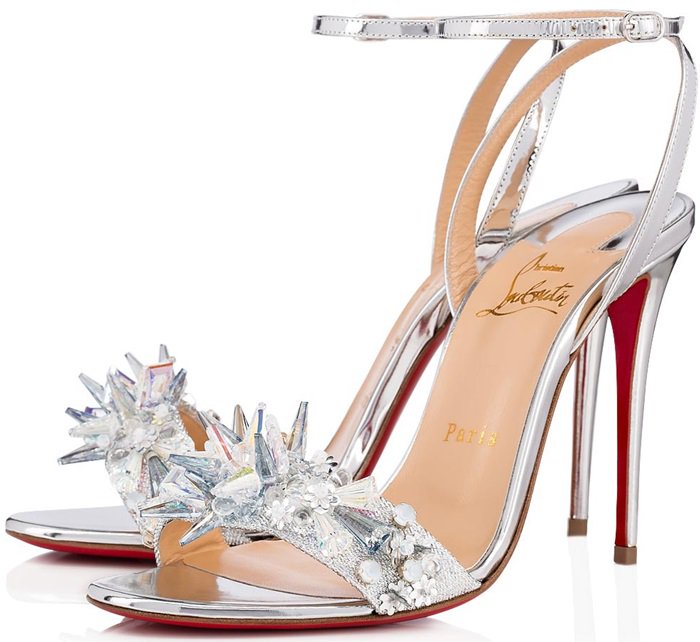 A starburst of glinting crystals, glimmering stones and sequins shines at the vamp of a silvery ankle strap sandal set on a slender stiletto heel