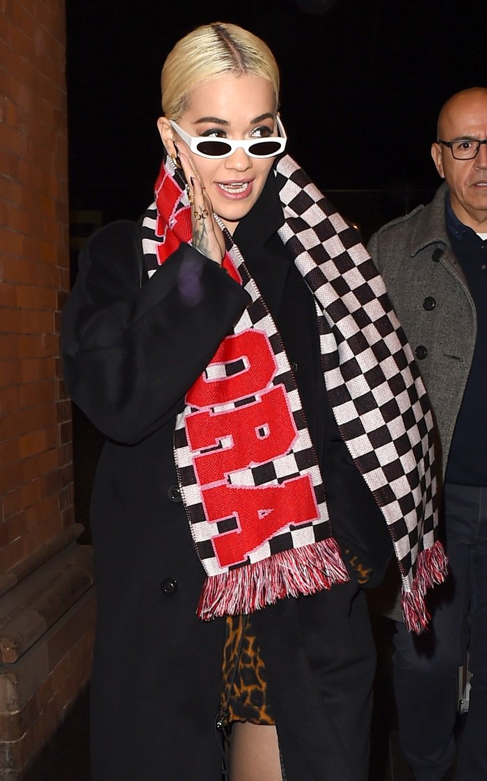 Rita Ora keeps warm in a Max Mara double-breasted wool coat, adding a bold touch with her House of Holland checkerboard scarf, animal-print mini dress, and white cat-eye Nor sunglasses while arriving at St Pancras railway station in London