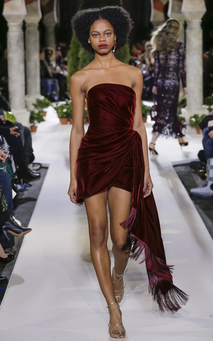Oscar de la Renta’s Fall 2019 runway showcases the deep-burgundy strapless velvet mini dress with dramatic draping and a fringed asymmetric hem, styled with delicate metallic strappy heels