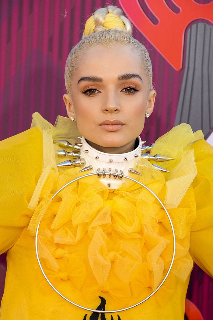 Poppy elevates her avant-garde look with a dramatic white choker adorned with silver spikes and a bold oversized metal ring pendant, perfectly complementing her ruffled yellow ensemble