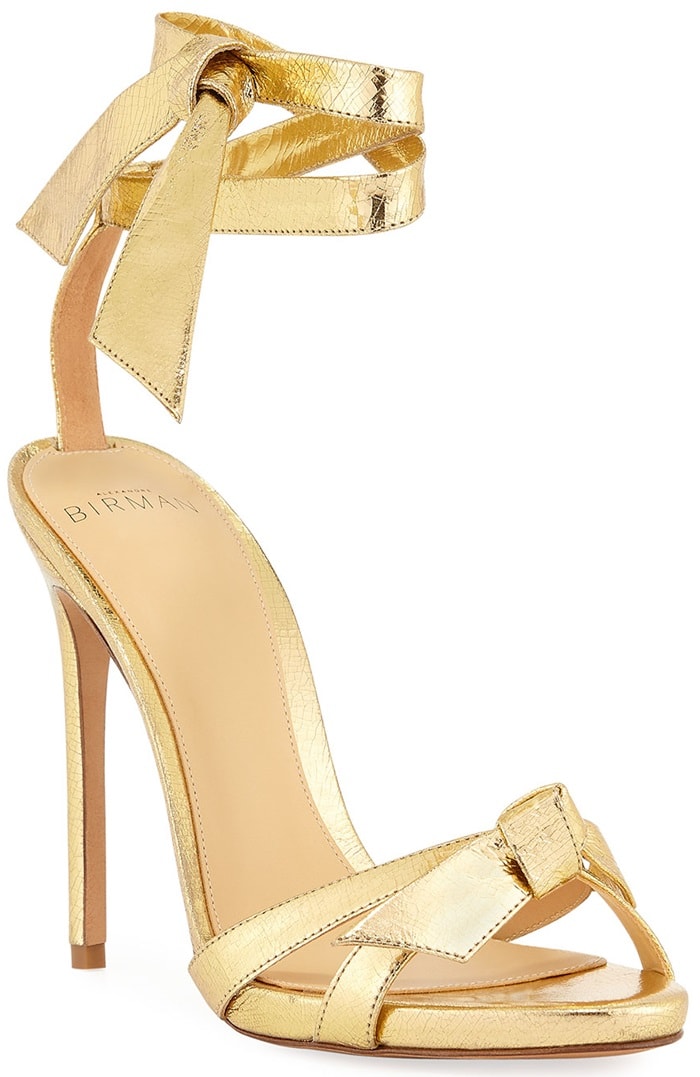 Alexandre Birman’s Clarita sandals in crackled metallic gold leather, defined by the signature hand-knotted bow at the toe and elegant ankle-wrap ties set on a slender stiletto heel