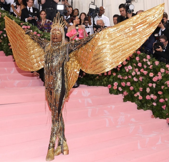 Spreading metallic wings across the Met Gala staircase, Billy Porter transforms his gold sequined platforms into part of a full-scale performance piece rooted in camp and couture spectacle