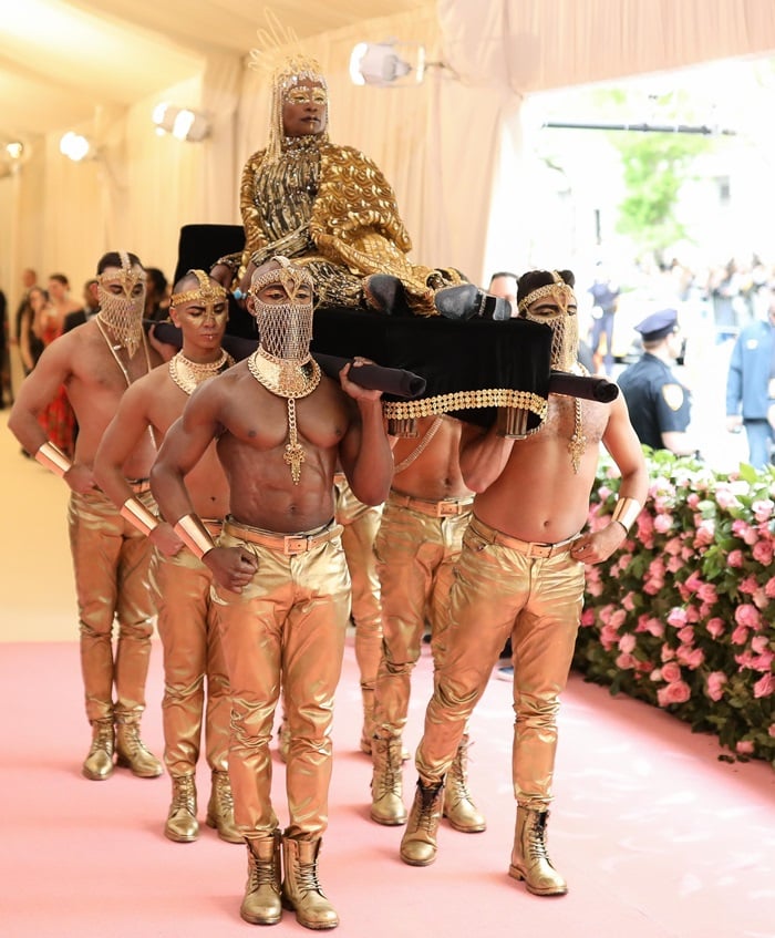 Billy Porter arrives at the 2019 Met Gala carried on a throne in towering gold platform boots and a radiant, winged “Sun God” ensemble inspired by ancient Egyptian royalty