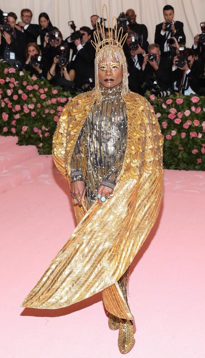 Billy Porter commands the Met Gala carpet in a crystal-drenched catsuit, sculptural gold cape, and glittering platform ankle boots that amplify the look’s theatrical grandeur
