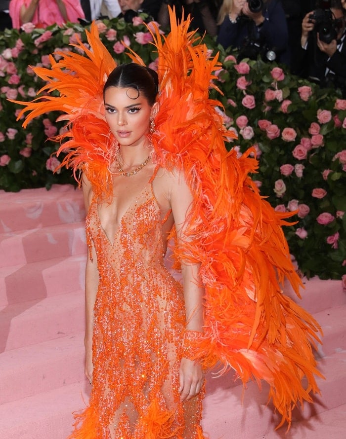 Kendall Jenner turned heads in a fiery orange Versace gown inspired by Cher, featuring sheer paneling, sparkling embellishments, and an explosion of feathers that playfully drew “chicken dress” comparisons online