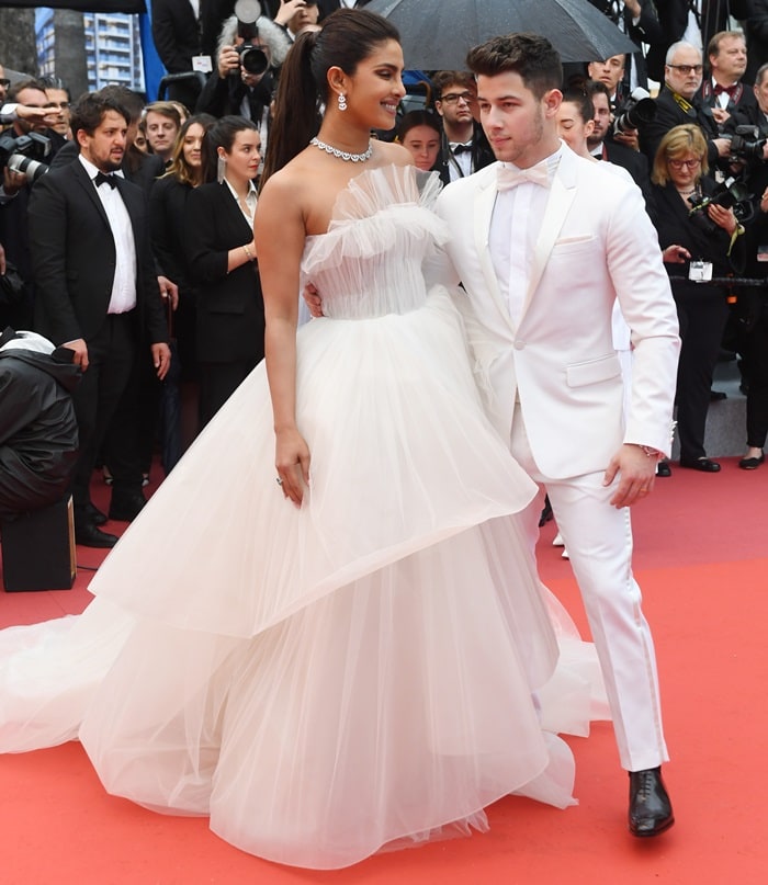 Priyanka Chopra stuns in an off-white strapless tulle gown with cascading tiers, paired with hidden Le Silla heels for red-carpet stability, while Nick Jonas complements her bridal-inspired look in a crisp white tuxedo and polished black patent leather shoes at the 2019 Cannes Film Festival
