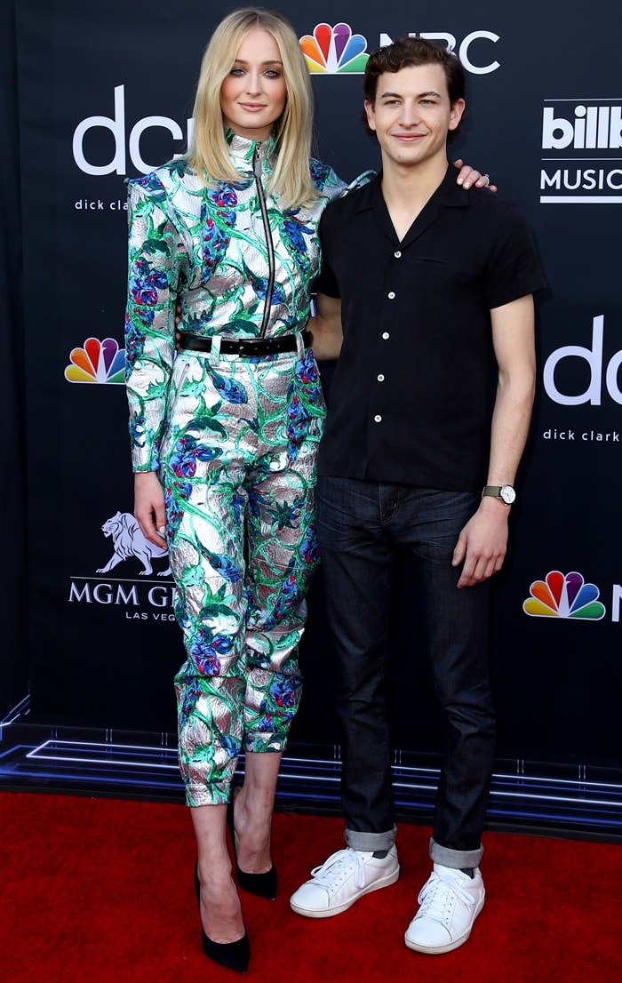 At the 2019 Billboard Music Awards in Las Vegas, Sophie Turner towered over Tye Sheridan in a metallic Louis Vuitton Asian Flower jacquard top and matching pants, grounding the futuristic look with classic black Jimmy Choo “Anouk” pumps, while he kept it casual in a Basic Rights shirt, Double 11 jeans, and white sneakers