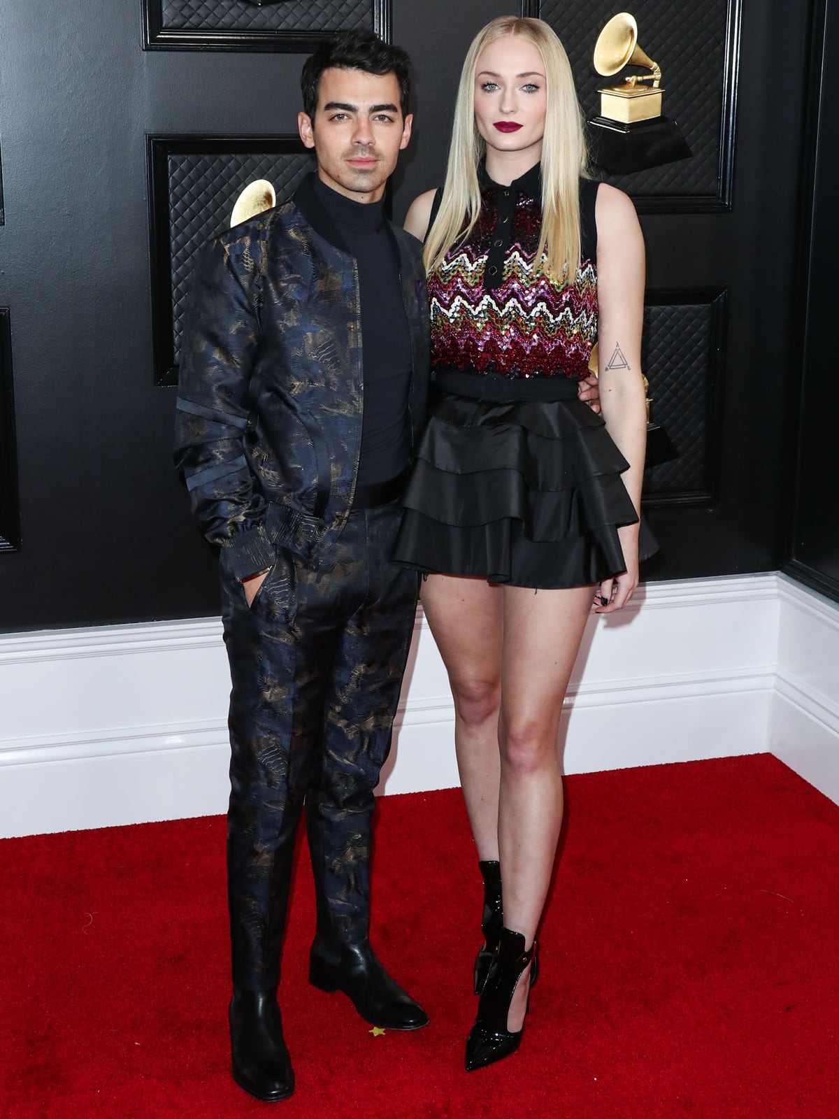 Sophie Turner towers in black patent Louis Vuitton ankle-strap heels paired with a ruffled mini dress, coordinating with Joe Jonas in a dark Ermenegildo Zegna suit at the 2020 Grammys red carpet