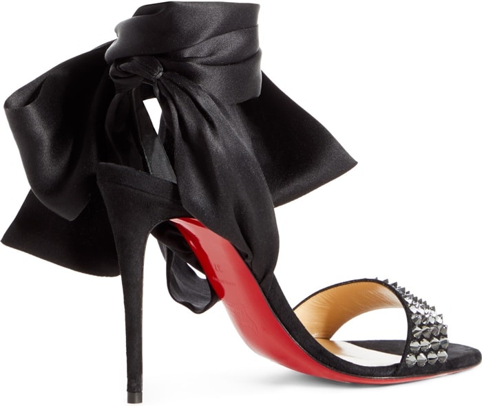 The Christian Louboutin Krystal Du Desert sandal is shown in isolation, highlighting its 100mm stiletto heel, matte black suede finish, and oversized satin ties set against the house’s iconic red lacquered sole