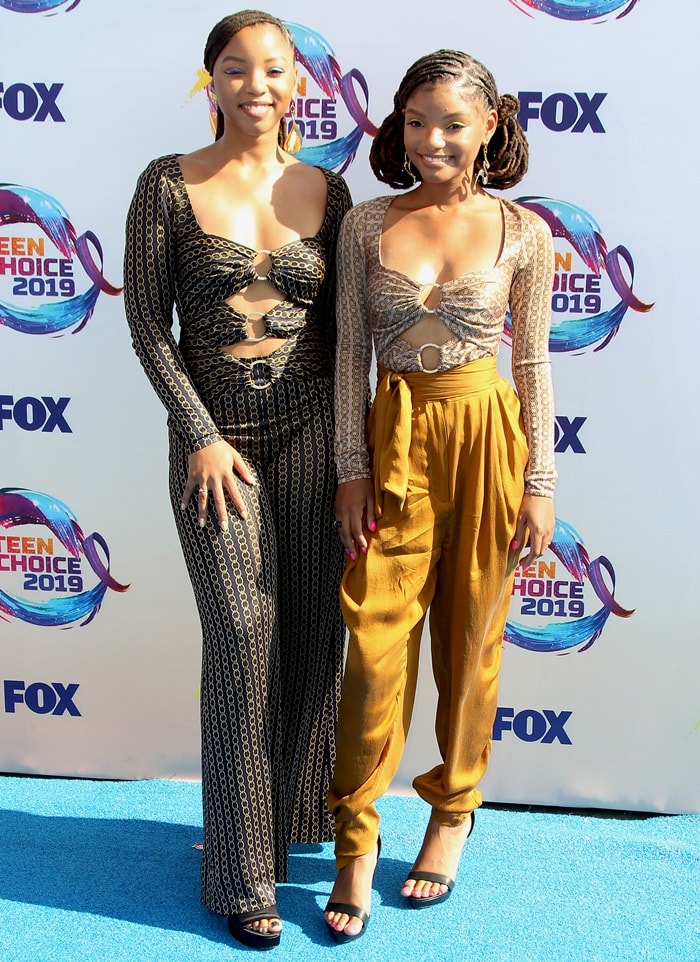 Chloe x Halle arrive at the 2019 Teen Choice Awards in coordinated Jonathan Simkhai chain-jacquard cutout bodysuits, with Chloe styling hers with matching wide-leg trousers and Halle pairing hers with Zimmermann Resistance high-waisted silk tapered pants and black Aldo Madalene platform sandals