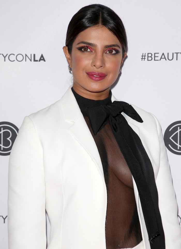 Vivid pink-and-black eye makeup echoed the event's beauty-focused energy, while a dramatic Christopher Bu pussy-bow blouse added contrast beneath the crisp white blazer