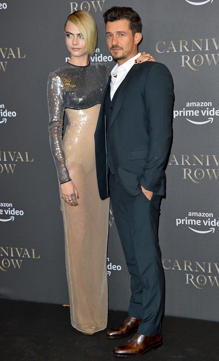 Cara Delevingne poses with Orlando Bloom in a sequined David Koma Resort 2020 gown, with her metallic platform heels just visible beneath the floor-length hem at the Carnival Row premiere in Berlin