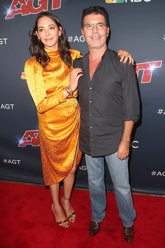 Lauren Silverman poses alongside Simon Cowell on the America’s Got Talent red carpet, wearing a marigold silk dress paired with nude Gianvito Rossi slingback sandals