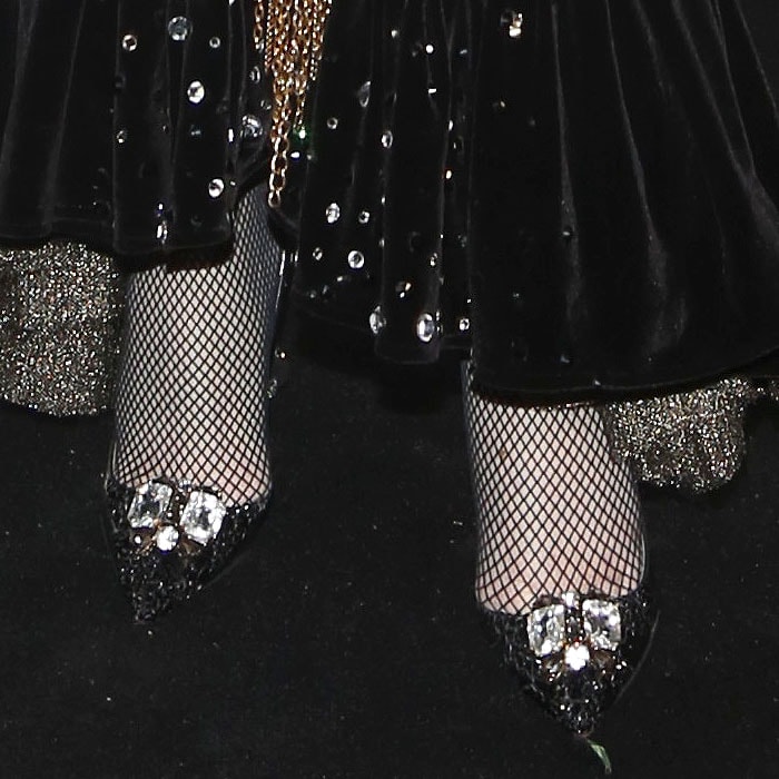 Her fishnet-clad feet step forward in custom crystal-encrusted Christian Louboutin pumps that mirror the gown’s glittering accents