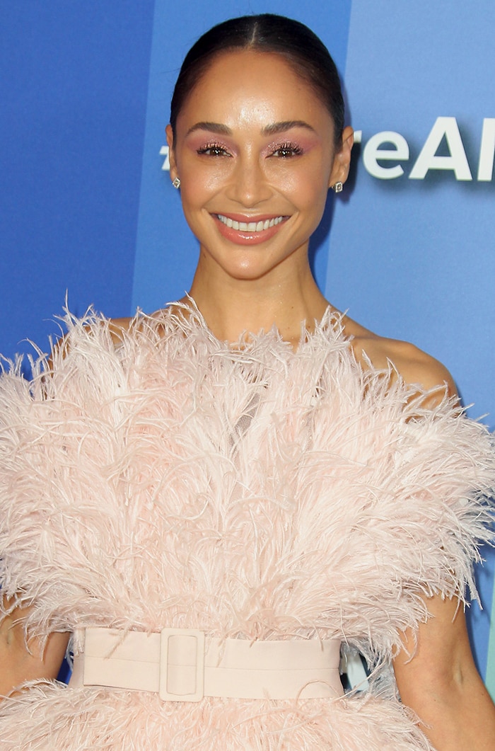 Cara Santana radiates with soft pink eyeshadow and glossy lips, her sleek hair pulled back into a polished low bun, accessorized with delicate stud earrings that complete her chic, feminine look