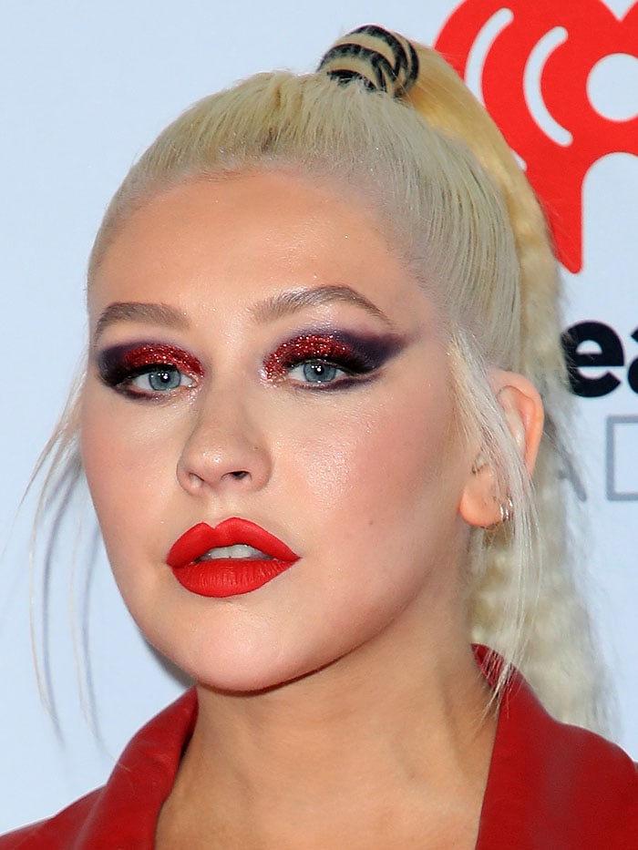 Her beauty look completes the statement with a crimped high ponytail, glitter-saturated red eyeshadow, and sharply defined lips that echo the outfit’s theatrical intensity