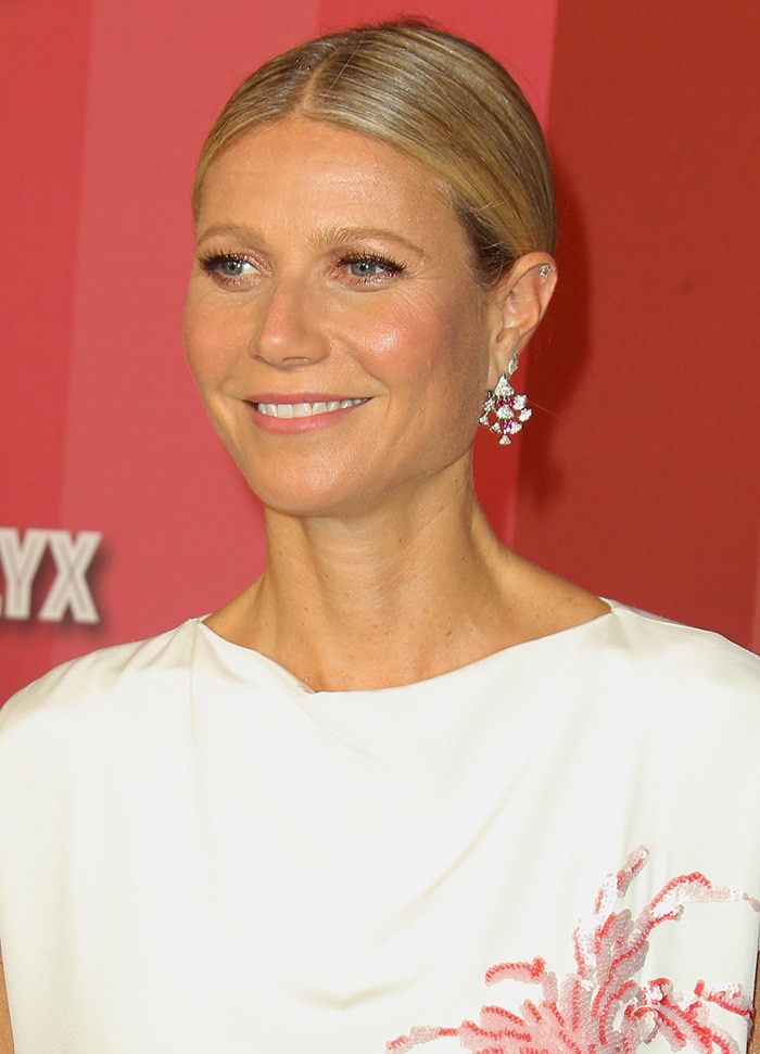 Gwyneth Paltrow’s hair is styled in a sleek, center-parted low bun, complemented by radiant, natural makeup with soft pink lips