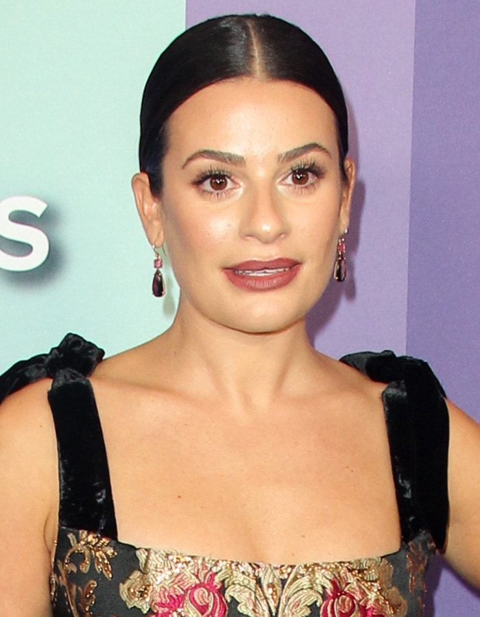 Lea Michele's hair is styled in a sleek, center-parted low bun, complemented by bold lashes, a deep berry lip, and statement drop earrings