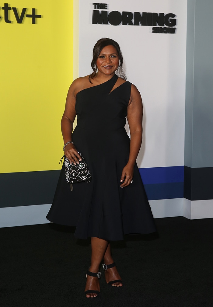 Mindy Kaling steps onto The Morning Show black carpet in a sculptural one-shoulder black dress, grounding the flared silhouette with elegant ankle-strap sandals that add just the right amount of sparkle and lift