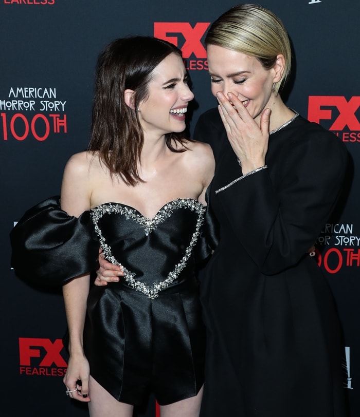 Sarah Paulson and Emma Roberts share a candid, lighthearted moment, with Paulson’s structured black dress—finished with a crystal-trimmed, heart-shaped neckline and voluminous off-the-shoulder sleeves—adding a sculptural, noir-inspired edge alongside Roberts’ more streamlined, minimalist silhouette