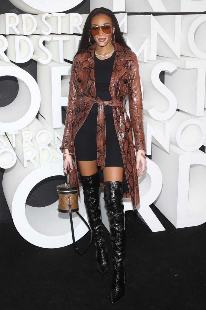 Winnie Harlow wearing a snakeskin trench coat, black croc-embossed thigh-high boots, and a Louis Vuitton Cannes handbag