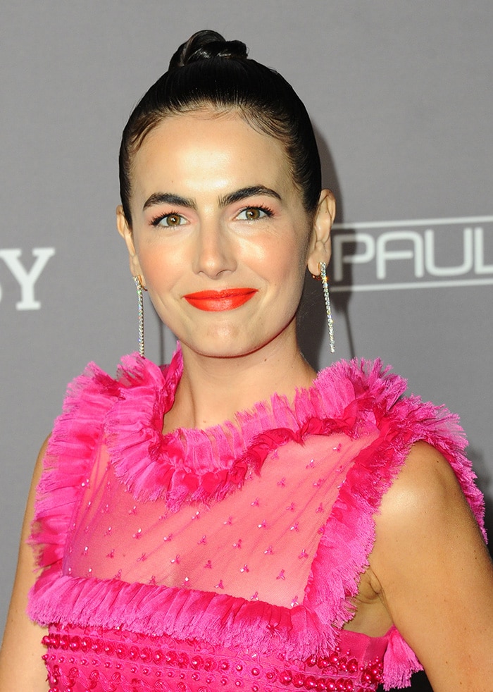 Camilla Belle pulls her hair into a bun and wears an orange-red lip shade