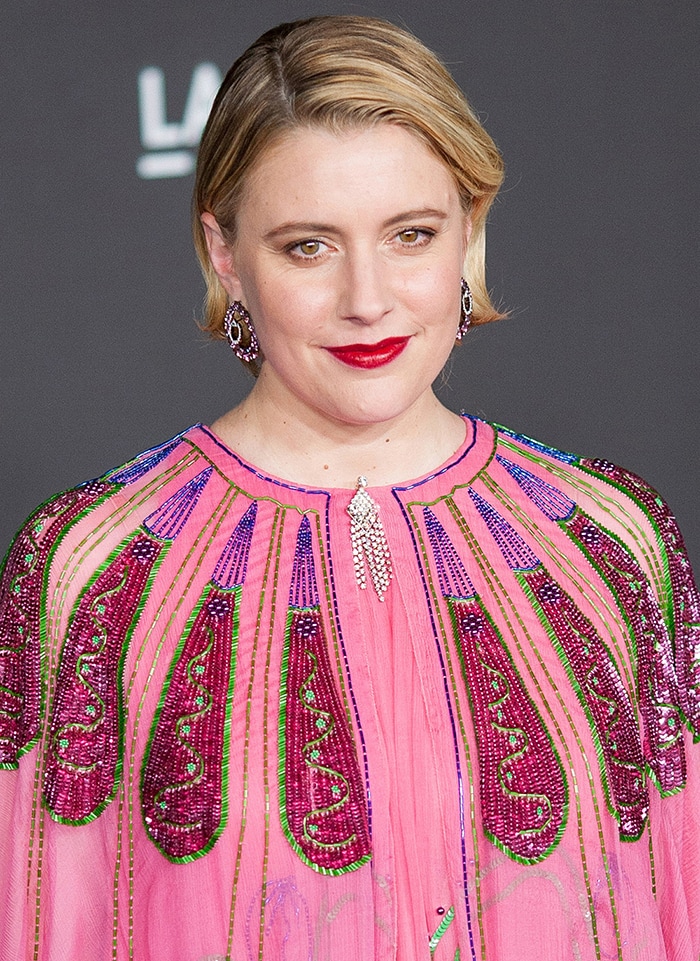 With a sleek side-part and bold berry lip, Greta Gerwig adds refined contrast to the gown’s vibrant, bohemian embellishment