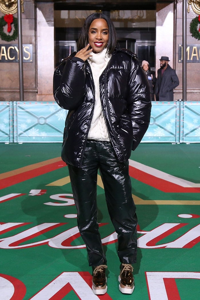 Kelly Rowland layered a glossy black puffer jacket and matching textured pants over a chunky white cable-knit sweater, grounding the oversized winter look with metallic gold Stella McCartney “Elyse” platform sneakers outside Macy’s Herald Square