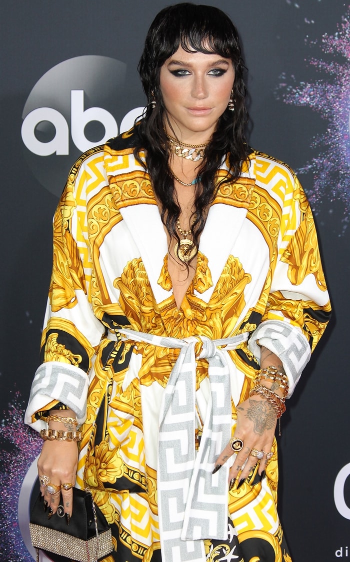 Kesha layers chunky gold jewelry over a plunging Versace robe featuring the house’s signature baroque motifs at the 2019 American Music Awards