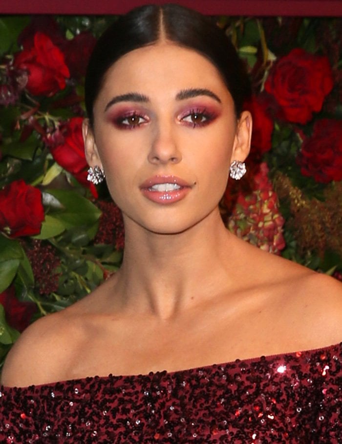Naomi Scott complements her outfit with red smoky eyes, glossy lips, and a sleek center-parted ponytail