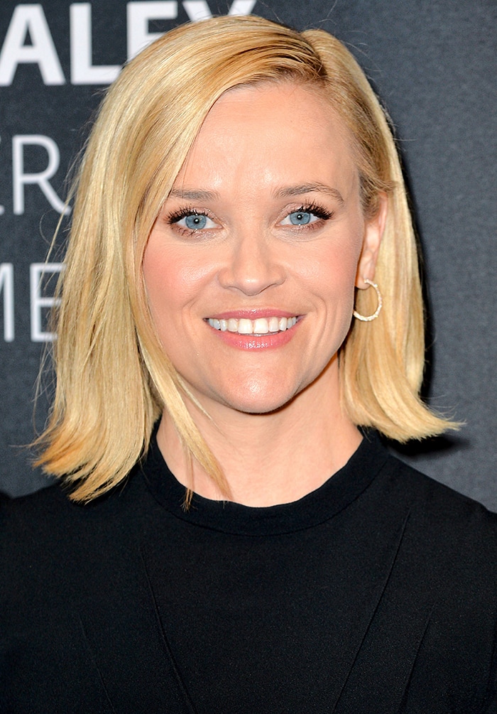 A close-up reveals Reese’s soft pink makeup, side-parted blonde bob, and understated gold hoops for a polished red-carpet finish