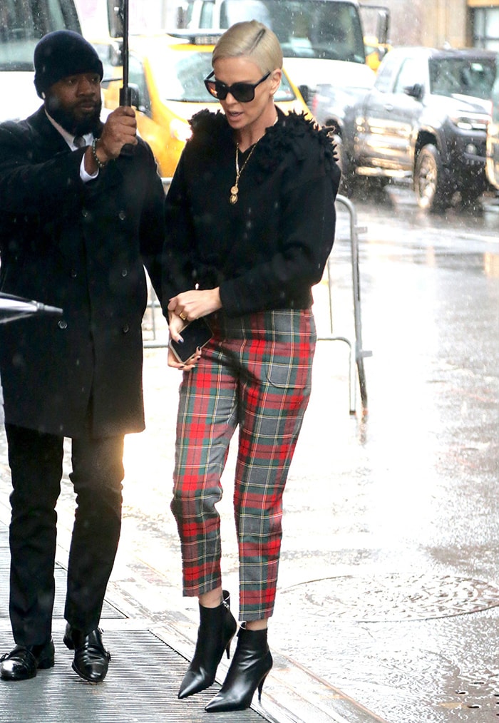 Charlize Theron sharpens her rainy-day street style with cropped plaid trousers and sleek black pointed-toe ankle boots, balancing classic tailoring with a sharp stiletto finish
