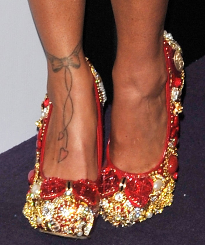 Katie Price highlights her bow-and-hearts foot tattoo in ornate red and gold embellished pumps, with crystal detailing and sculptural accents drawing the eye to the delicate ink along her ankle and instep